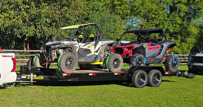 Black 24 ft flatbed trailer hitched to a white pickup in a grassy yard, carrying two Polaris RZR side-by-sides secured at the wheels with green wheel nets and blue ratchet straps