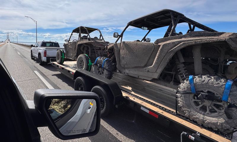 View from a following vehicle of a white pickup towing the 24 ft flatbed with two muddy side-by-sides strapped down at the wheels, crossing the Mackinac Bridge