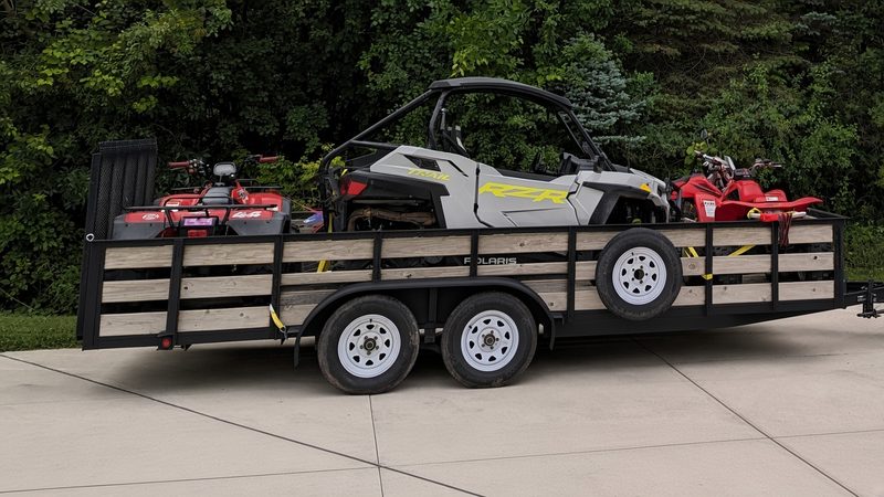 Black-framed 18 ft tandem-axle landscape trailer with natural wood side rails and a spare tire, loaded with two red ATVs and a grey Polaris RZR side-by-side, parked on a concrete driveway
